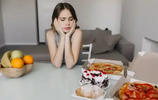 Mujer con ansiedad por comer frente a pizza y tarta en la mesa, con fruta al lado