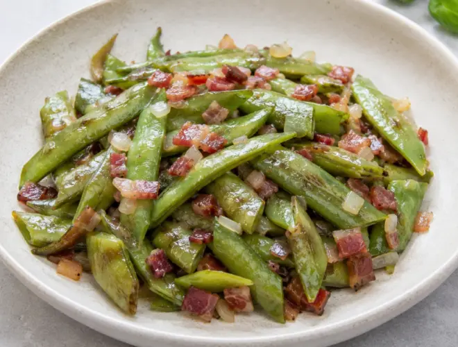 Habas salteadas con jam&oacute;n serrano y cebolleta, servidas en plato blanco, foto gourmet luminosa con fondo neutro.