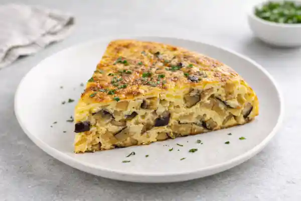 Tortilla de berenjenas jugosa con dados de berenjena y cebolla, servida en plato blanco con luz natural y fondo neutro