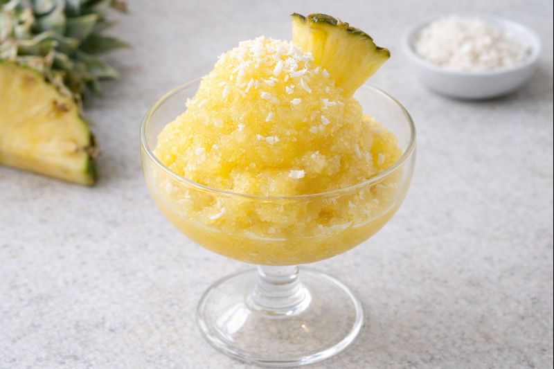 Sorbete de pi&ntilde;a y coco en copa de cristal, textura granizada y cremosa, con virutas de coco y gajo de pi&ntilde;a sobre fondo neutro luminoso