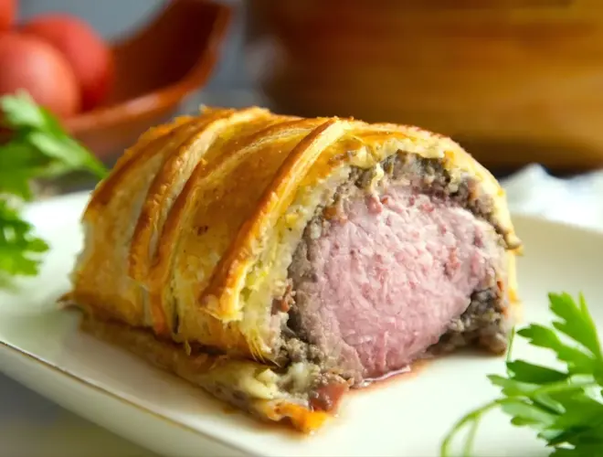 Solomillo Wellington de ternera envuelto en hojaldre dorado, cortado en rebanadas y servido en plato, con cebolla caramelizada.